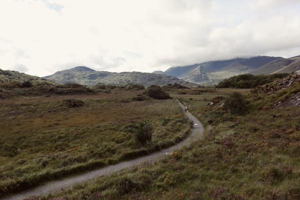 A winding dirt path stretches through a vast, green valley, flanked by rolling hills and distant mountains shrouded in clouds